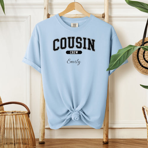 Chambray light blue Comfort Colors T-shirt featuring a bold “Cousin Crew” design in black varsity-style lettering, with the name “Your Name” in elegant script below. Displayed knotted at the bottom and hanging on a wooden hanger in a cozy, neutral-toned room with natural decor elements.