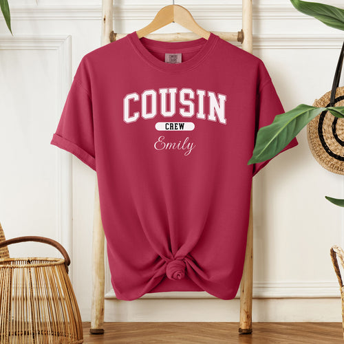 Chili Comfort Colors T-shirt featuring a bold “Cousin Crew” design in white varsity-style lettering, with the name “Your Name” in elegant script below. Displayed knotted at the bottom and hanging on a wooden hanger in a cozy, neutral-toned room with natural decor elements.