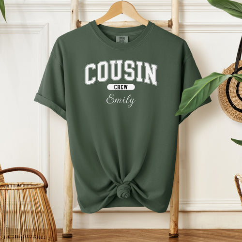 Hemp color Comfort Colors T-shirt featuring a bold “Cousin Crew” design in white varsity-style lettering, with the name “Your Name” in elegant script below. Displayed knotted at the bottom and hanging on a wooden hanger in a cozy, neutral-toned room with natural decor elements.