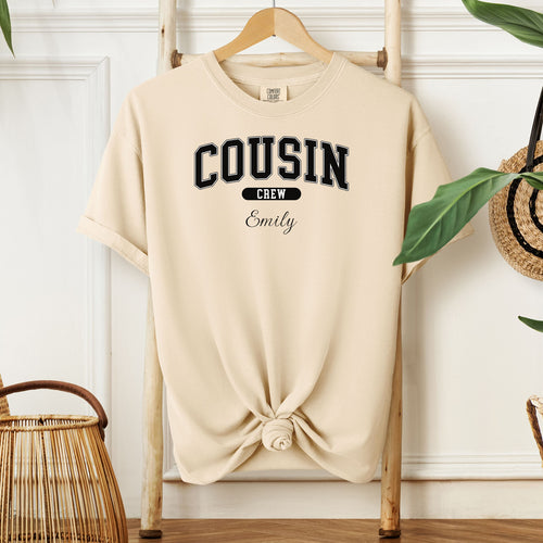 Ivory Comfort Colors T-shirt featuring a bold “Cousin Crew” design in black varsity-style lettering, with the name “Your Name” in elegant script below. Displayed knotted at the bottom and hanging on a wooden hanger in a cozy, neutral-toned room with natural decor elements.