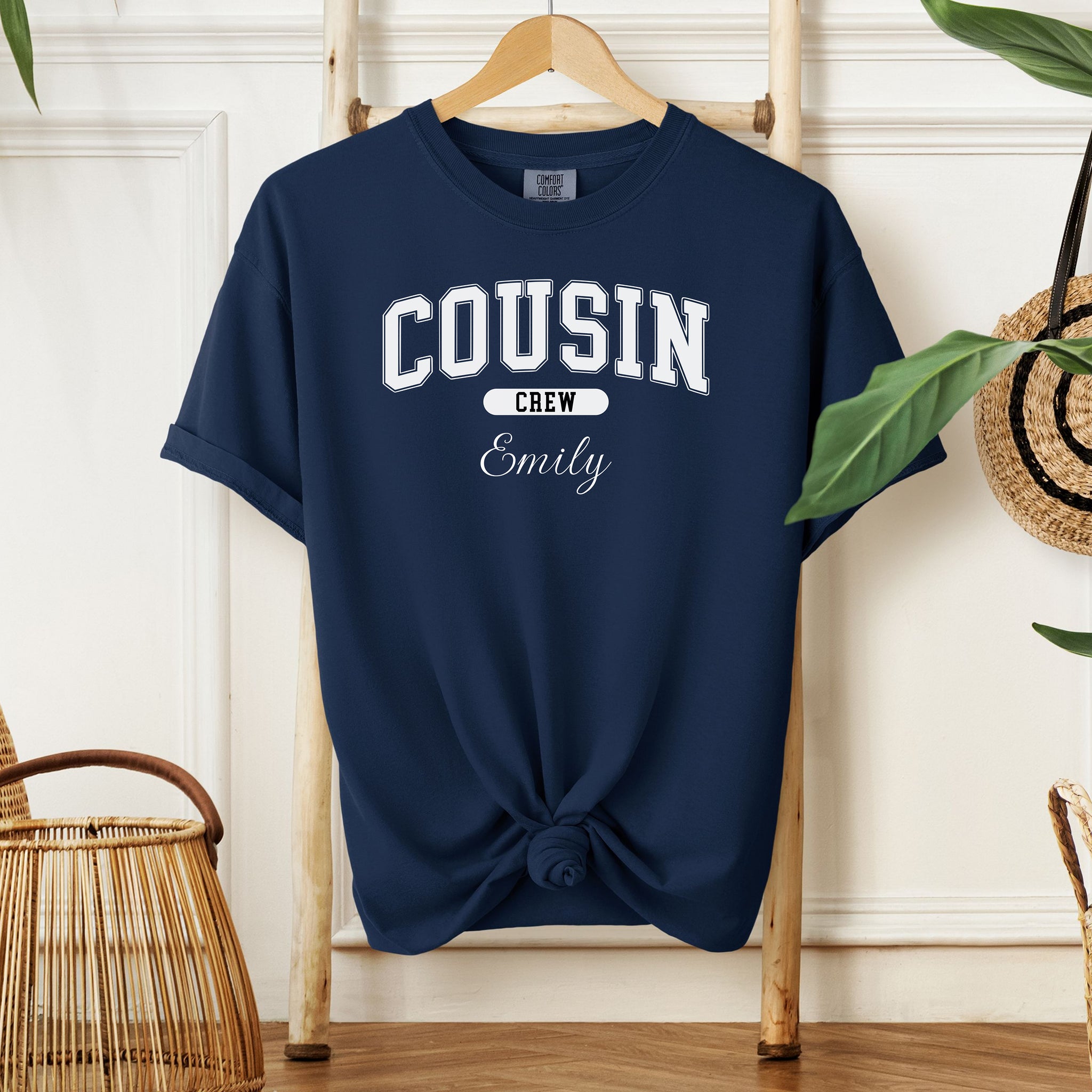 True Navy Comfort Colors T-shirt featuring a bold “Cousin Crew” design in white varsity-style lettering, with the name “Your Name” in elegant script below. Displayed knotted at the bottom and hanging on a wooden hanger in a cozy, neutral-toned room with natural decor elements.