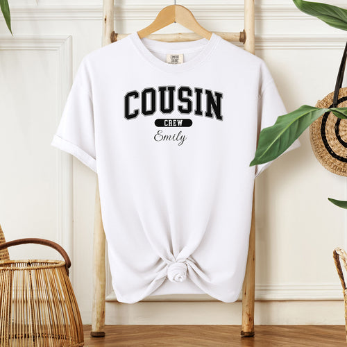 White Comfort Colors T-shirt featuring a bold “Cousin Crew” design in black varsity-style lettering, with the name “Your Name” in elegant script below. Displayed knotted at the bottom and hanging on a wooden hanger in a cozy, neutral-toned room with natural decor elements.