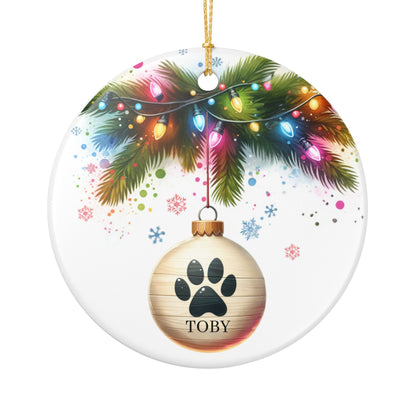 Personalized ceramic Christmas ornament with paw print and pet's name, surrounded by festive Christmas lights and tree branches.