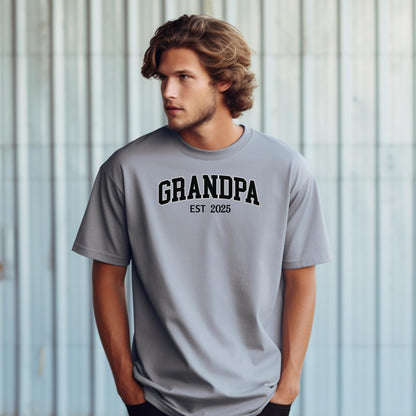 Granite t-shirt featuring a bold collegiate-style 'GRANDPA EST [Year]' design in black and white lettering. Perfect gift for new grandfathers to celebrate the year they became a grandpa. A thoughtful, casual, and comfortable shirt ideal for Father's Day, family gatherings, or everyday wear.