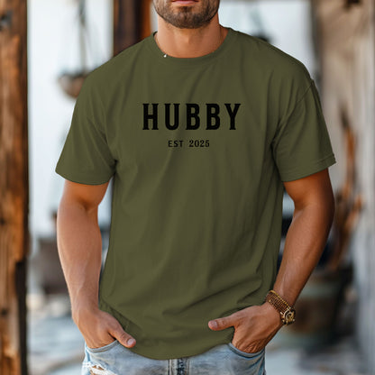 Hubby Est [Year] design printed on a Hemp color unisex t-shirt. The shirt features bold black capital letters spelling 'HUBBY' with 'EST [Year]' in smaller text below. Perfect for newlyweds, anniversaries, or wedding gifts, this casual and modern design celebrates marriage in a simple, stylish way. 