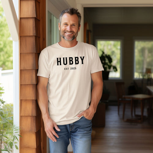 Hubby Est [Year] design printed on a Ivory unisex t-shirt. The shirt features bold black capital letters spelling 'HUBBY' with 'EST [Year]' in smaller text below. Perfect for newlyweds, anniversaries, or wedding gifts, this casual and modern design celebrates marriage in a simple, stylish way. 
