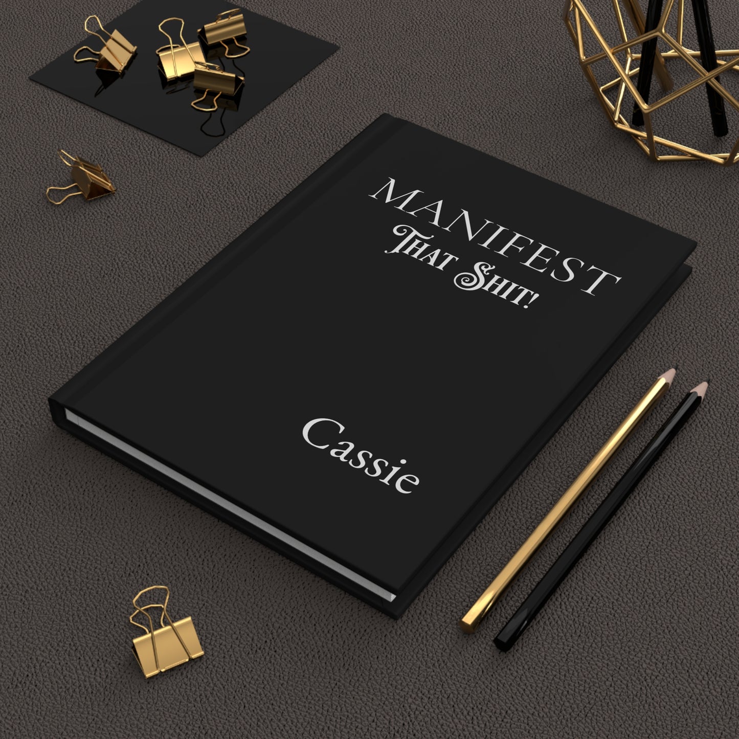 Hardcover journal with 'Manifest That Shit!' and name on a dark surface with office supplies.