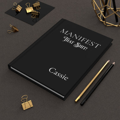 Hardcover journal with 'Manifest That Shit!' and name on a dark surface with office supplies.