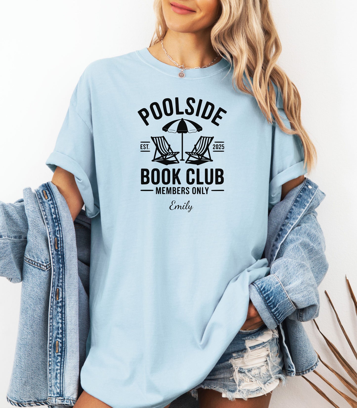 Person wearing a Chambray light blue T-shirt featuring a "Poolside Book Club Members Only" design with two lounge chairs and an umbrella graphic, personalized with the name you choose.