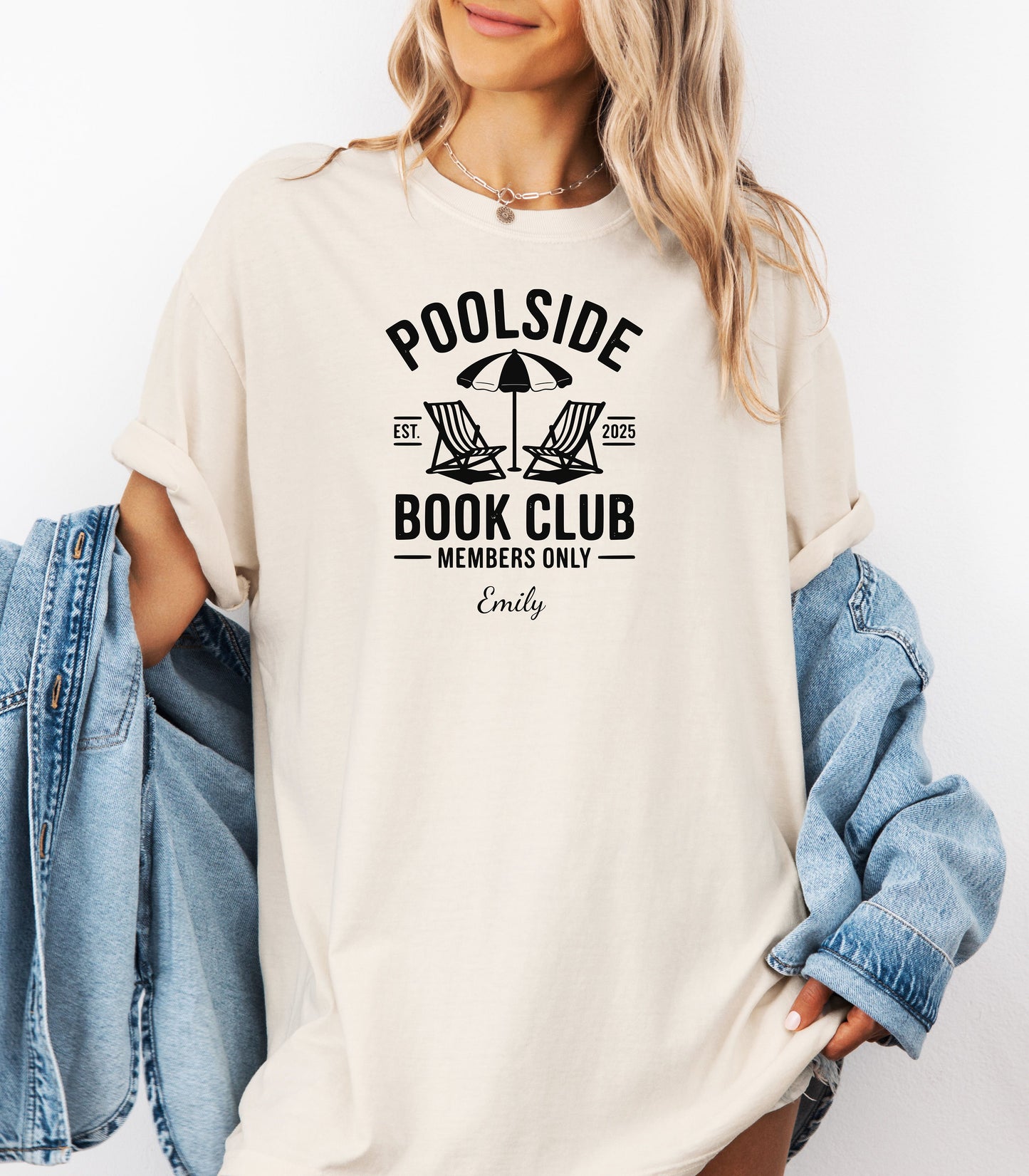 Person wearing an Ivory T-shirt featuring a "Poolside Book Club Members Only" design with two lounge chairs and an umbrella graphic, personalized with the name you choose.