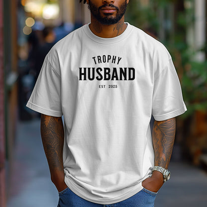 White Trophy Husband EST [Year] graphic T-shirt featuring bold black typography on a casual crewneck shirt. Perfect gift for newlyweds, anniversaries, grooms, or husbands with a sense of humor. Comfortable, stylish, and great for everyday wear or special occasions to celebrate marriage and fun couple vibes.