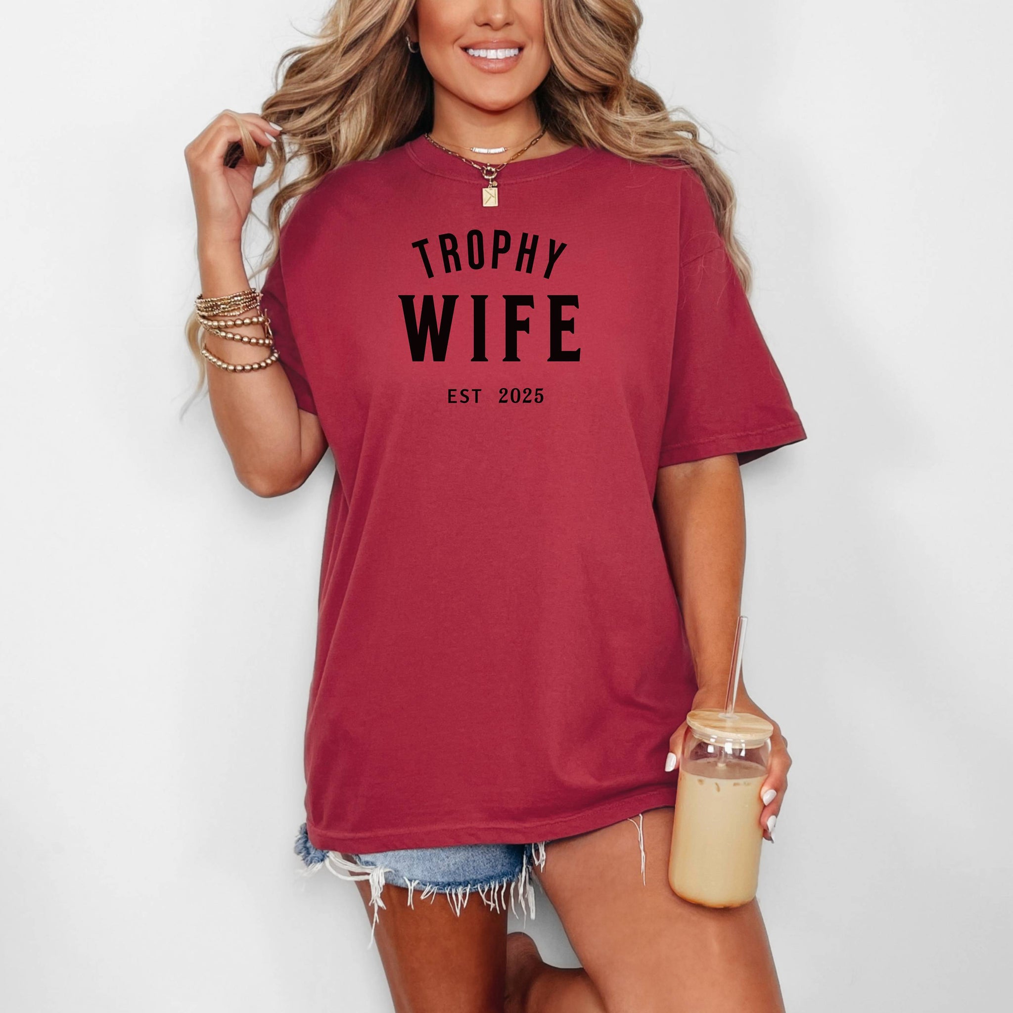 "Trophy Wife Est [Year] design on a chili unisex t-shirt. The shirt features bold black lettering with 'TROPHY WIFE' in large uppercase letters and 'EST [Year]' below in smaller text. A fun and stylish design perfect for newlyweds, anniversaries, or bridal gifts, celebrating marriage with a confident modern look. Modeled in a casual, relaxed fit style."