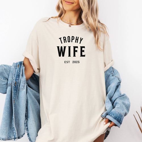 "Trophy Wife Est [Year] design on an Ivory unisex t-shirt. The shirt features bold black lettering with 'TROPHY WIFE' in large uppercase letters and 'EST [Year]' below in smaller text. A fun and stylish design perfect for newlyweds, anniversaries, or bridal gifts, celebrating marriage with a confident modern look. Modeled in a casual, relaxed fit style."
