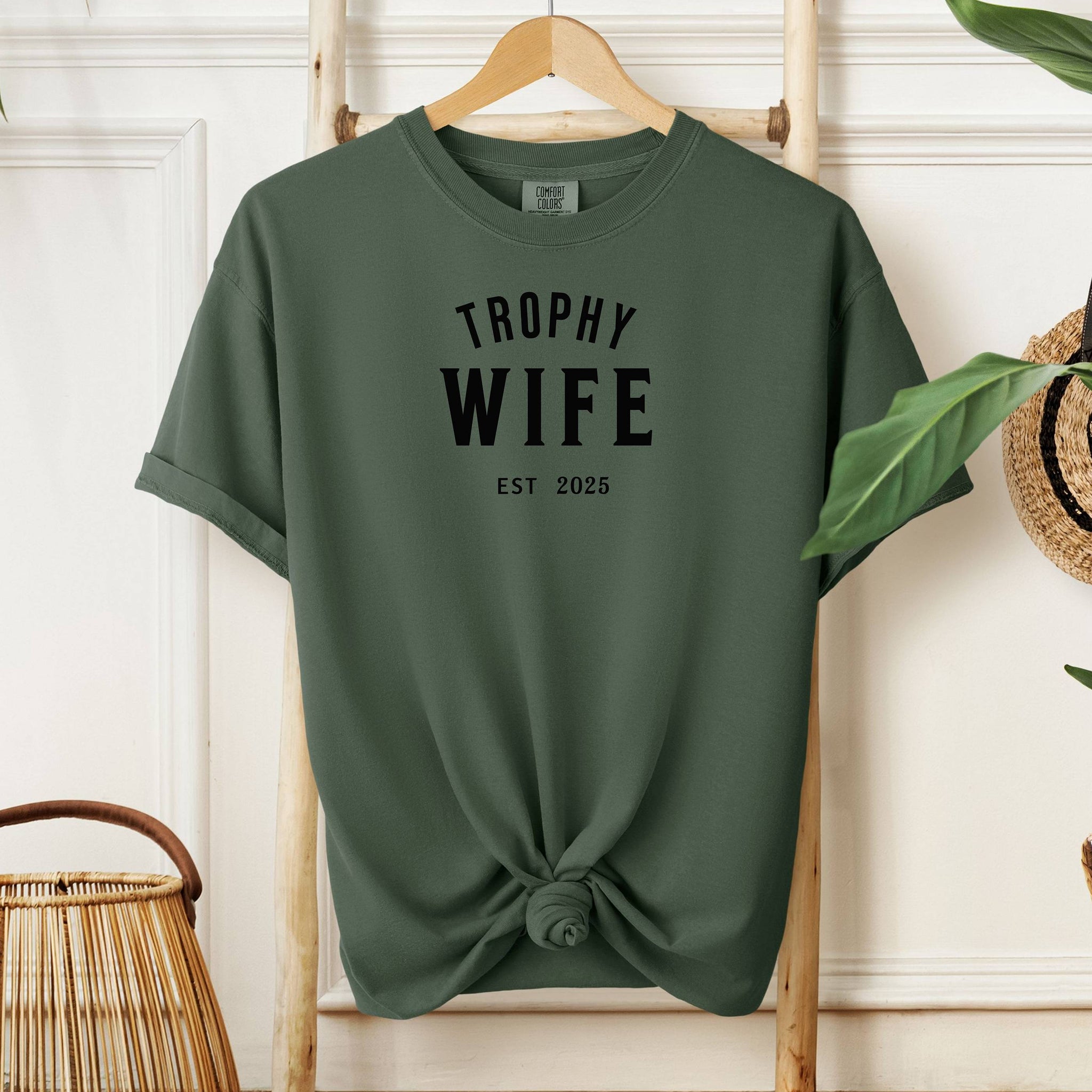 "Trophy Wife Est [Year] design on a moss unisex t-shirt. The shirt features bold black lettering with 'TROPHY WIFE' in large uppercase letters and 'EST [Year]' below in smaller text. A fun and stylish design perfect for newlyweds, anniversaries, or bridal gifts, celebrating marriage with a confident modern look. Modeled in a casual, relaxed fit style."