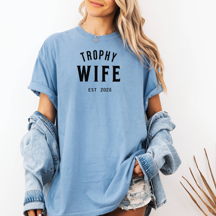 "Trophy Wife Est [Year] design on a washed denim unisex t-shirt. The shirt features bold black lettering with 'TROPHY WIFE' in large uppercase letters and 'EST [Year]' below in smaller text. A fun and stylish design perfect for newlyweds, anniversaries, or bridal gifts, celebrating marriage with a confident modern look. Modeled in a casual, relaxed fit style."