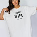 "Trophy Wife Est [Year] design on a white unisex t-shirt. The shirt features bold black lettering with 'TROPHY WIFE' in large uppercase letters and 'EST [Year]' below in smaller text. A fun and stylish design perfect for newlyweds, anniversaries, or bridal gifts, celebrating marriage with a confident modern look. Modeled in a casual, relaxed fit style."