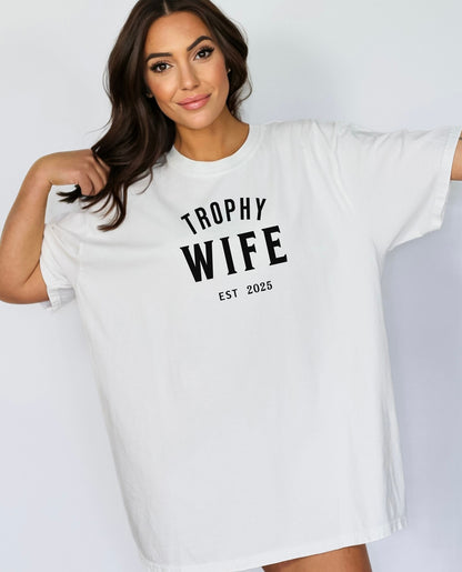 "Trophy Wife Est [Year] design on a white unisex t-shirt. The shirt features bold black lettering with 'TROPHY WIFE' in large uppercase letters and 'EST [Year]' below in smaller text. A fun and stylish design perfect for newlyweds, anniversaries, or bridal gifts, celebrating marriage with a confident modern look. Modeled in a casual, relaxed fit style."