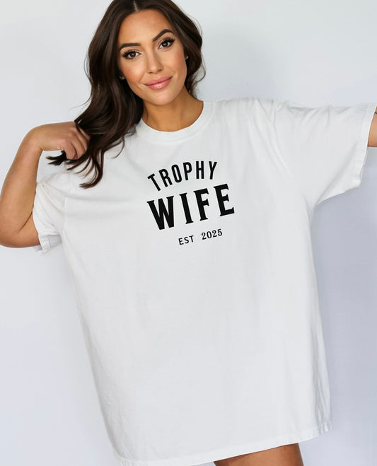 "Trophy Wife Est [Year] design on a white unisex t-shirt. The shirt features bold black lettering with 'TROPHY WIFE' in large uppercase letters and 'EST [Year]' below in smaller text. A fun and stylish design perfect for newlyweds, anniversaries, or bridal gifts, celebrating marriage with a confident modern look. Modeled in a casual, relaxed fit style."
