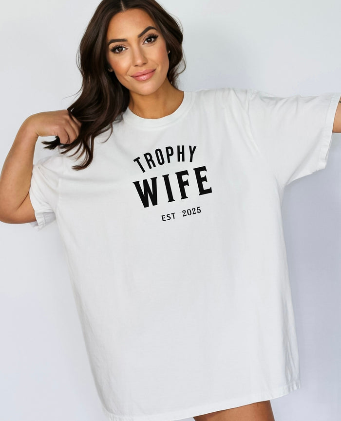 "Trophy Wife Est [Year] design on a white unisex t-shirt. The shirt features bold black lettering with 'TROPHY WIFE' in large uppercase letters and 'EST [Year]' below in smaller text. A fun and stylish design perfect for newlyweds, anniversaries, or bridal gifts, celebrating marriage with a confident modern look. Modeled in a casual, relaxed fit style."