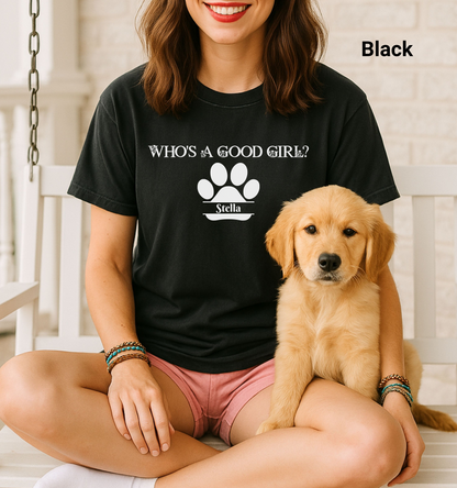 Woman wearing a black t-shirt that reads “Who’s a Good Girl?” above a white paw print graphic with the name “Stella” on a dog bone below. Sitting cross-legged with a golden retriever puppy, the shirt is a playful and heartwarming tribute to a beloved pet—perfect for dog moms and puppy lovers.
