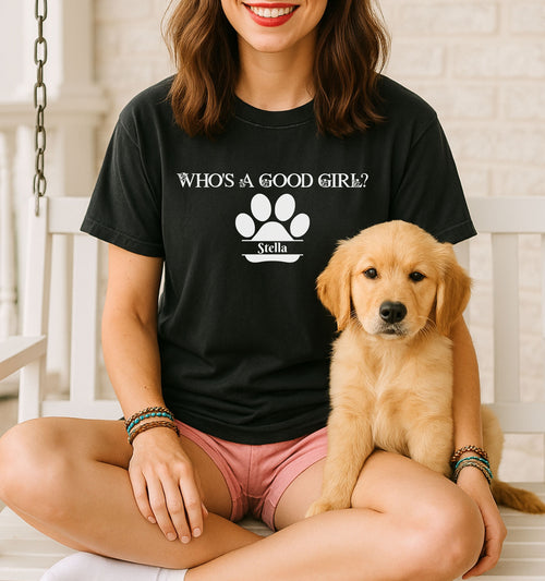 Woman wearing a black t-shirt that reads “Who’s a Good Girl?” above a white paw print graphic with the name “Stella”.   Sitting cross-legged with a golden retriever puppy, the shirt is a playful and heartwarming tribute to a beloved pet—perfect for dog moms and puppy lovers.