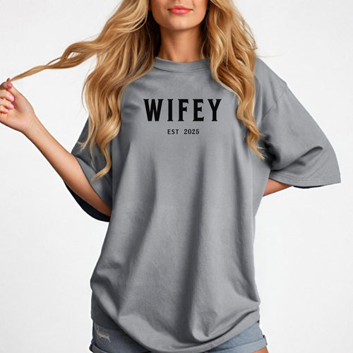 Granite T-shirt with the text "WIFEY EST 2025" in bold black lettering, celebrating a newlywed or engaged status. The modern minimalist design makes it perfect for brides, honeymoon outfits, or casual newlywed style. Displayed on a woman wearing denim shorts, holding her hair with a confident and playful pose, ideal for wedding gifts, bridal showers, or anniversary celebrations.