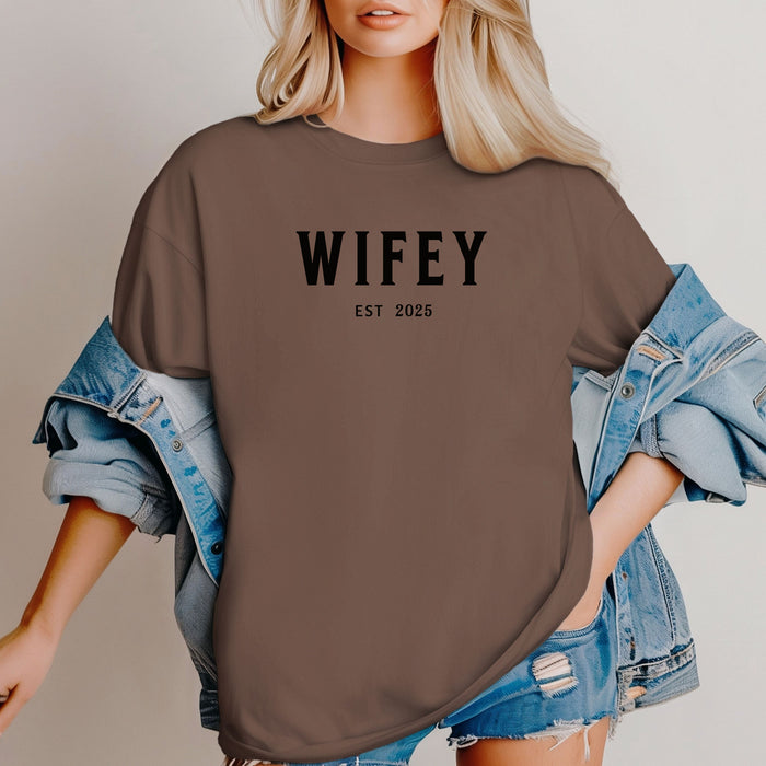 Espresso T-shirt with the text "WIFEY EST 2025" in bold black lettering, celebrating a newlywed or engaged status. The modern minimalist design makes it perfect for brides, honeymoon outfits, or casual newlywed style.  Ideal for wedding gifts, bridal showers, or anniversary celebrations.