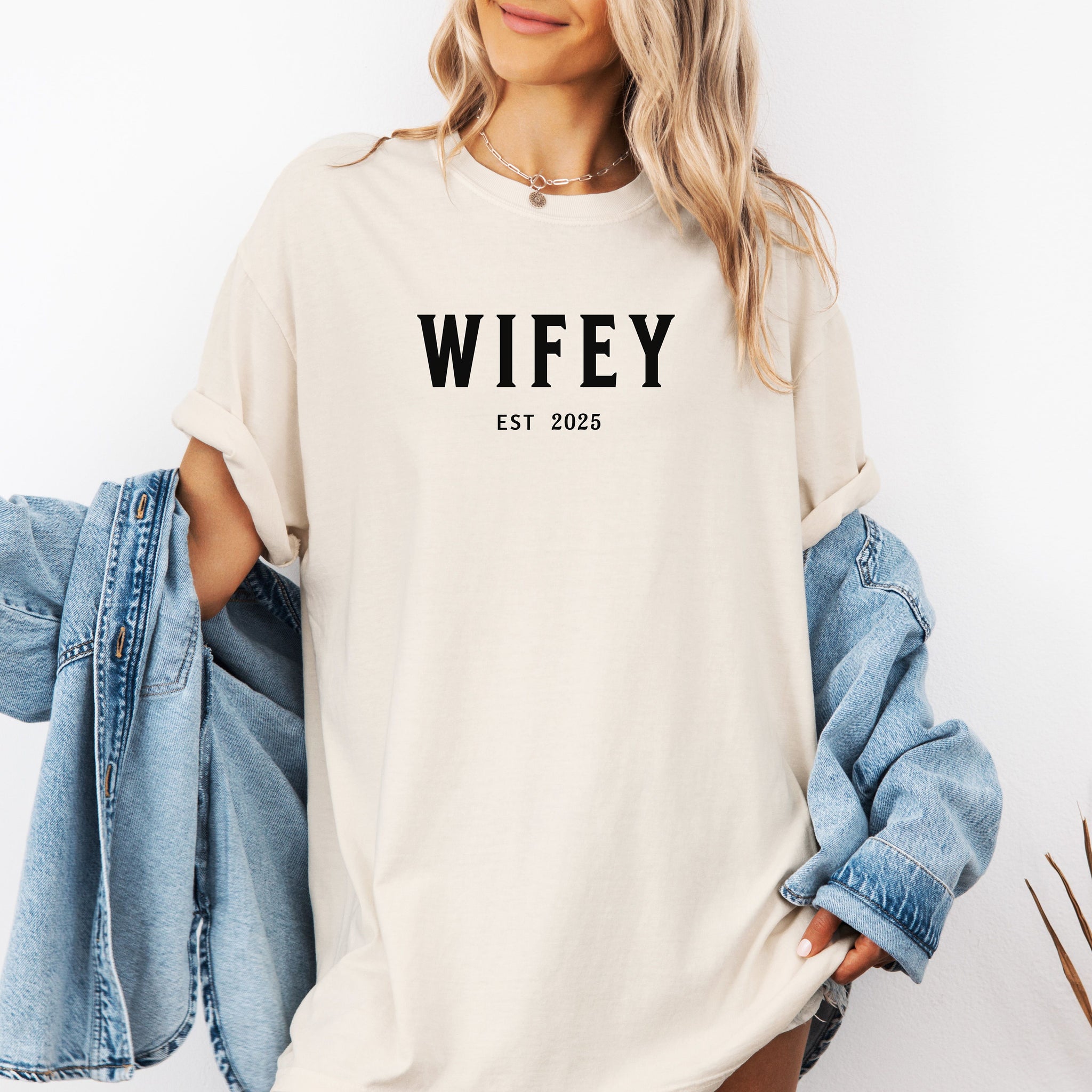 Ivory T-shirt with the text "WIFEY EST 2025" in bold black lettering, celebrating a newlywed or engaged status. The modern minimalist design makes it perfect for brides, honeymoon outfits, or casual newlywed style.  Ideal for wedding gifts, bridal showers, or anniversary celebrations.