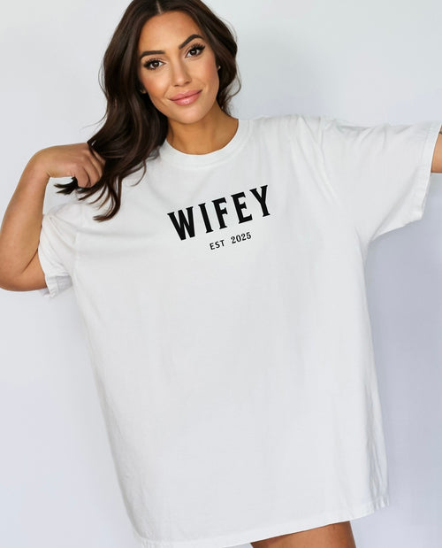 White T-shirt with the text "WIFEY EST 2025" in bold black lettering, celebrating a newlywed or engaged status. The modern minimalist design makes it perfect for brides, honeymoon outfits, or casual newlywed style.  Ideal for wedding gifts, bridal showers, or anniversary celebrations.