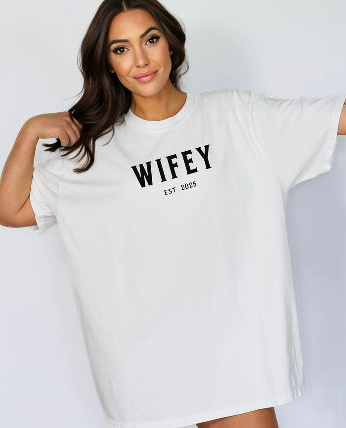 White T-shirt with the text "WIFEY EST 2025" in bold black lettering, celebrating a newlywed or engaged status. The modern minimalist design makes it perfect for brides, honeymoon outfits, or casual newlywed style.  Ideal for wedding gifts, bridal showers, or anniversary celebrations.
