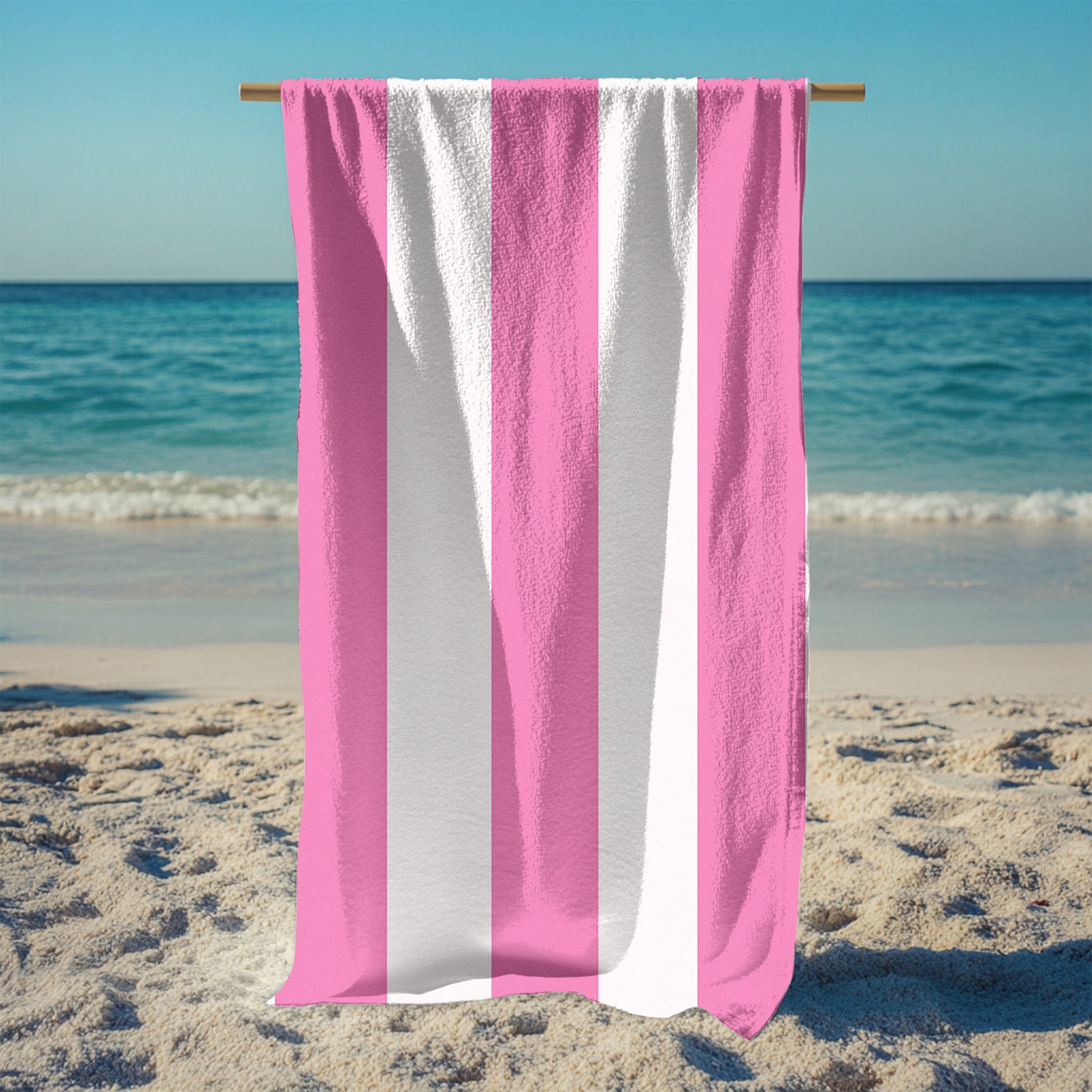 A standing holding up a beach towel with bold vertical pink and white stripes, standing on a sunny beach with the ocean in the background. The fresh, classic striped design brings a soft and stylish touch to any seaside escape, perfect for lounging, drying off, or making a beachside statement.