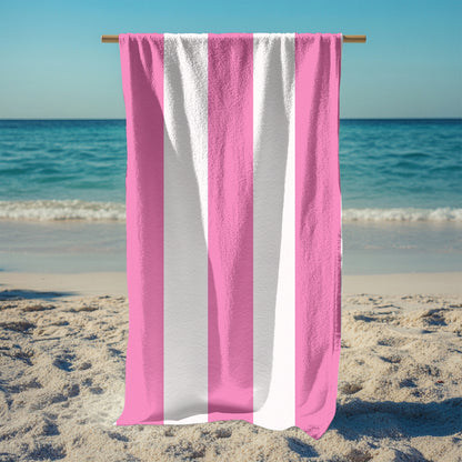 A standing holding up a beach towel with bold vertical pink and white stripes, standing on a sunny beach with the ocean in the background. The fresh, classic striped design brings a soft and stylish touch to any seaside escape, perfect for lounging, drying off, or making a beachside statement.