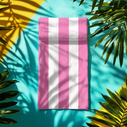 A beach towel with bold vertical pink and white stripes, folded neatly on colorful concrete.  The fresh, classic striped design brings a soft and stylish touch to any seaside escape, perfect for lounging, drying off, or making a beachside statement.