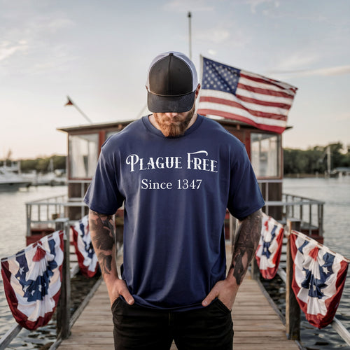 Person wearing a True Navy T-shirt with a humorous Renaissance Faire design that reads “Plague Free Since 1347” in medieval-style lettering, celebrating fair and festival fun with a nod to history. A playful shirt perfect for history buffs, medieval enthusiasts, and anyone attending a Renaissance fair or themed event.