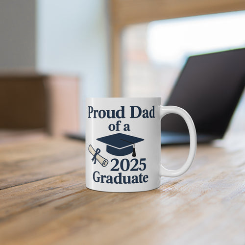 White mug with 'Proud Dad of a 2025 Graduate' text, a diploma, and a graduation cap sitting on a desk with a computer in the background.