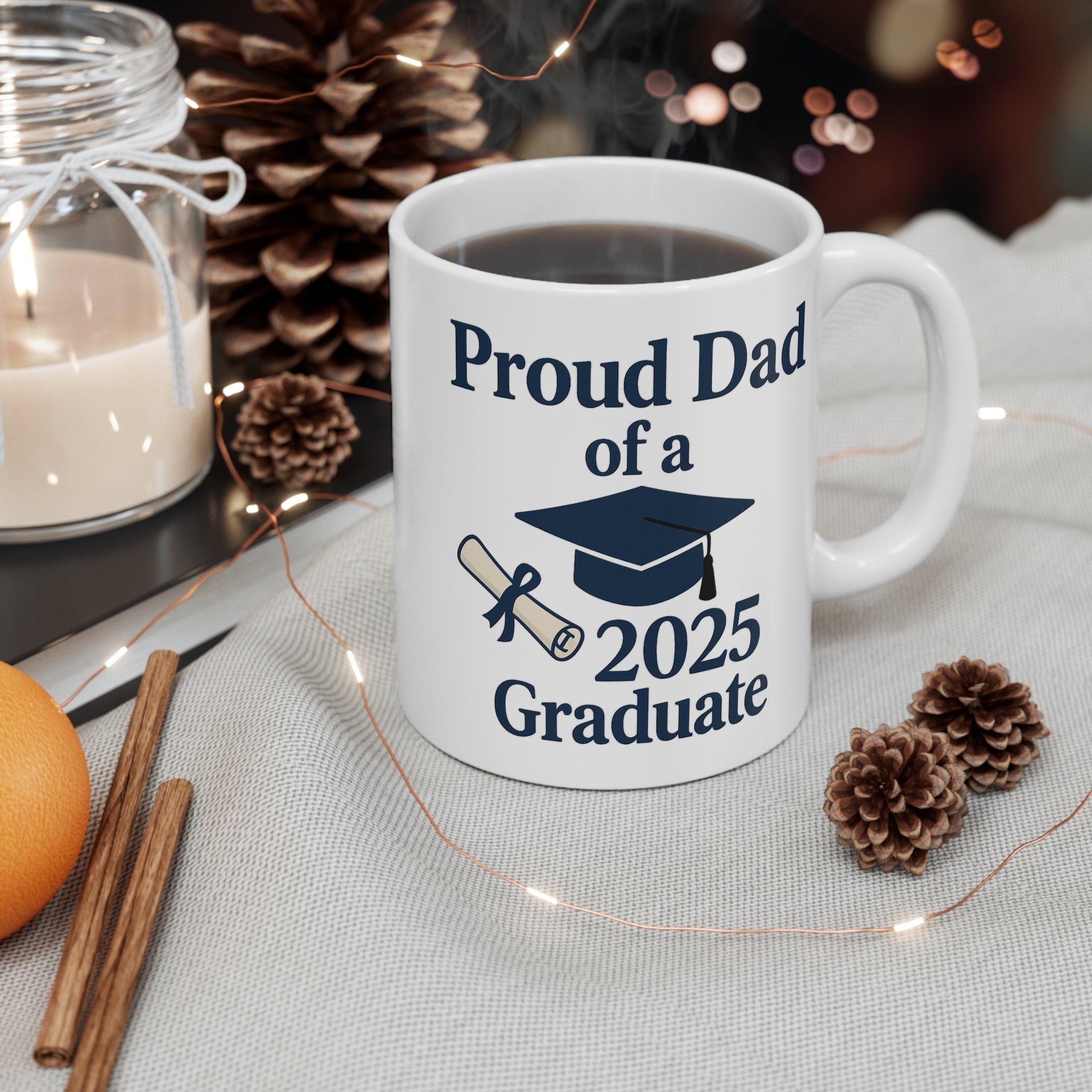 A white mug with 'Proud Dad of a 2025 Graduate' text, a diploma, and a graduation cap sitting on a table with fall, holiday decor and a candle around it.  The cup is filled with steaming hot coffee.