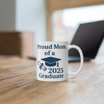 A white mug with 'Proud Mom of a 2025 Graduate' text, a diploma, and a graduation cap sitting on a desk with a computer behind it.