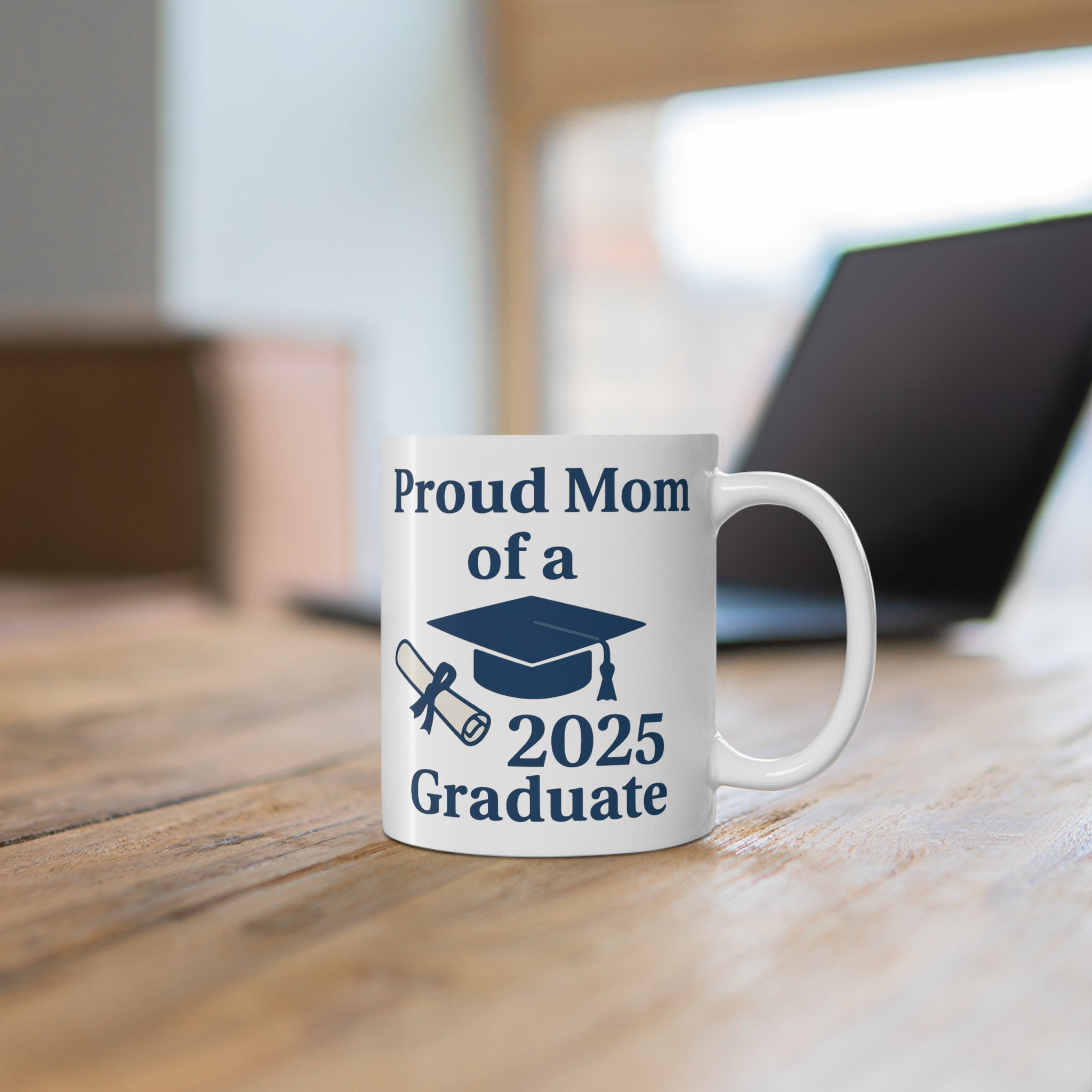 A white mug with 'Proud Mom of a 2025 Graduate' text, a diploma, and a graduation cap sitting on a desk with a computer behind it.