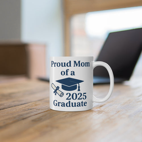 A white mug with 'Proud Mom of a 2025 Graduate' text, a diploma, and a graduation cap sitting on a desk with a computer behind it.