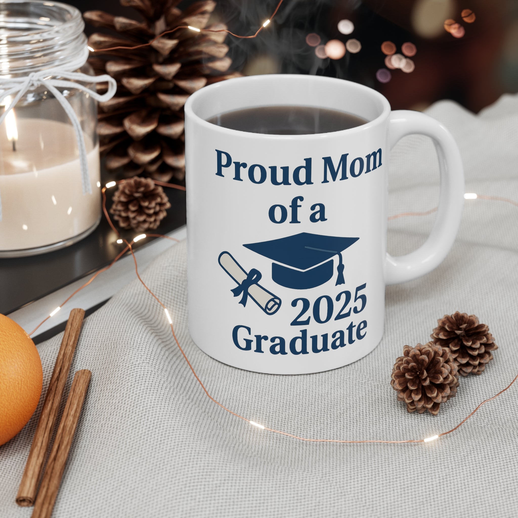A white mug with 'Proud Mom of a 2025 Graduate' text, a diploma, and a graduation cap sitting on a table with pinecones, a candle and holiday decor around it.