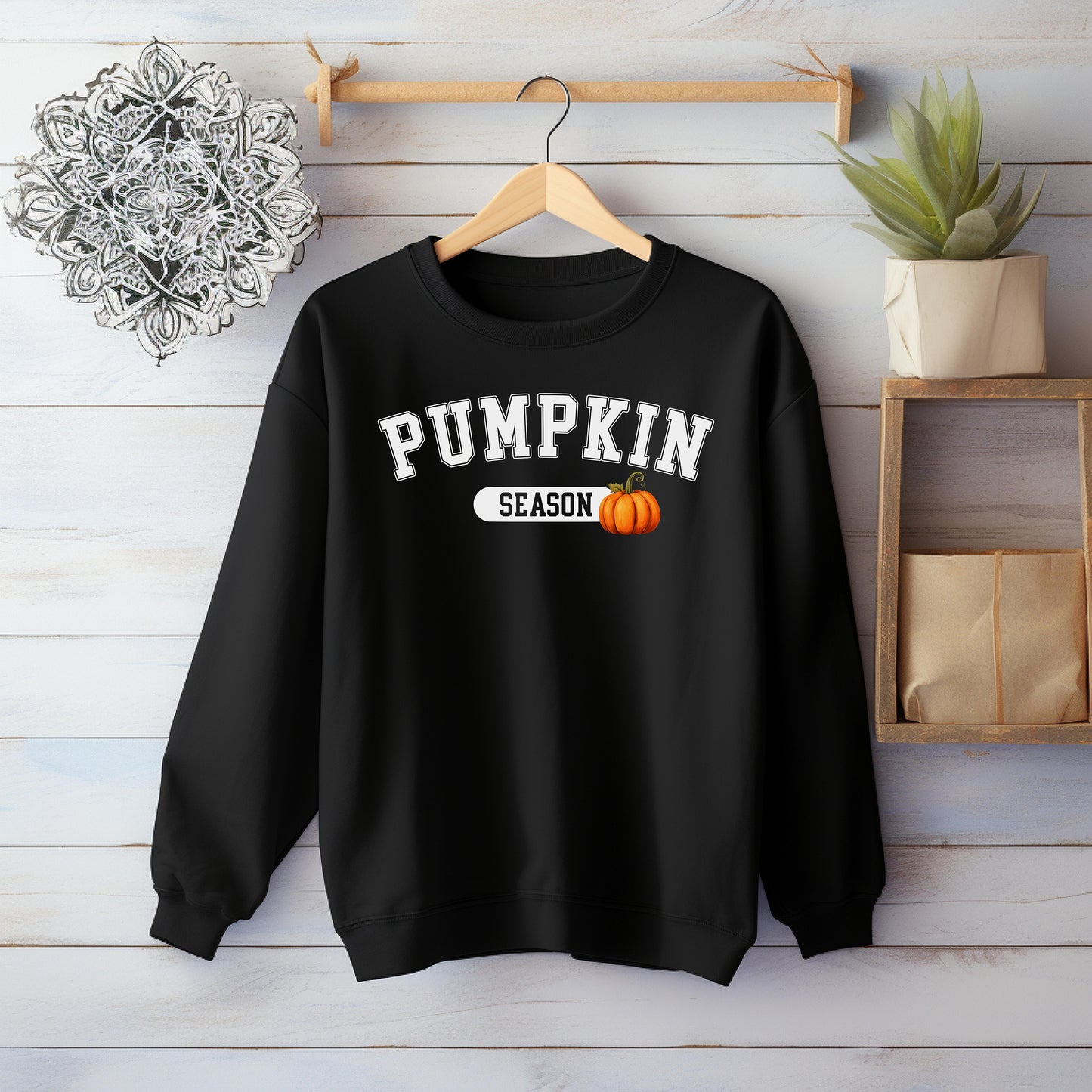 Black sweatshirt with 'PUMPKIN SEASON' text and pumpkin graphic on a wooden hanger against a wooden wall.
