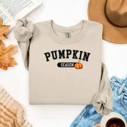 Sand sweatshirt with 'Pumpkin Season' text and pumpkin graphic on a white surface with autumn-themed items.
