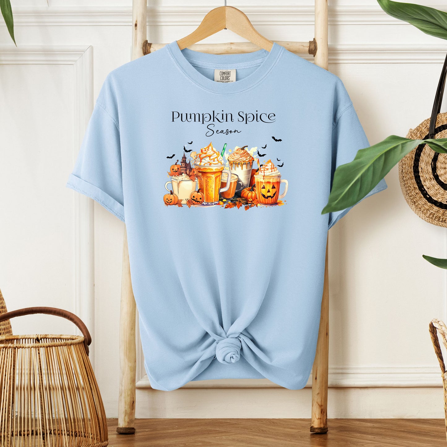 Chambray light blue T-shirt featuring a “Pumpkin Spice Season” design with colorful autumn drinks topped with whipped cream, surrounded by pumpkins, jack-o’-lanterns, fall leaves, and flying bats. The cozy, festive artwork celebrates the love for pumpkin spice and autumn vibes, perfect for seasonal wear and fall enthusiasts.