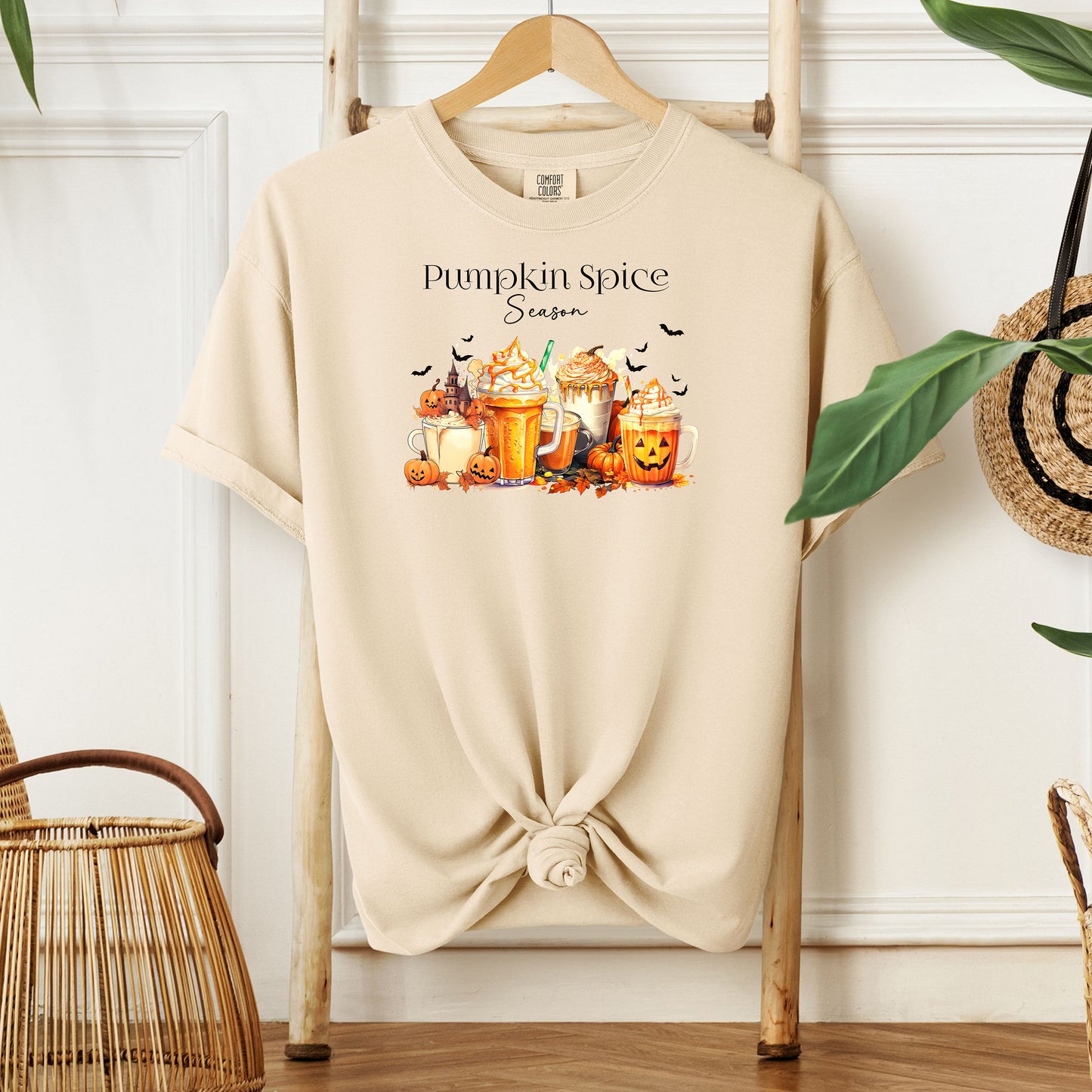 Ivory T-shirt featuring a “Pumpkin Spice Season” design with colorful autumn drinks topped with whipped cream, surrounded by pumpkins, jack-o’-lanterns, fall leaves, and flying bats. The cozy, festive artwork celebrates the love for pumpkin spice and autumn vibes, perfect for seasonal wear and fall enthusiasts.