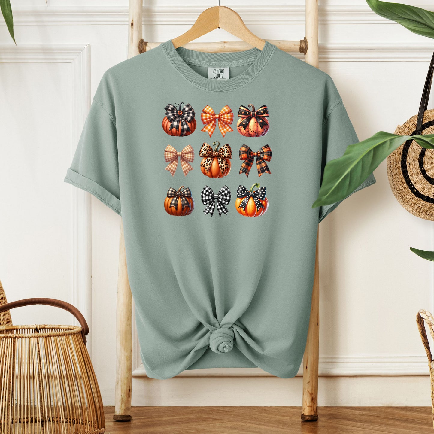 Bay T-shirt featuring “Pumpkins and Coquette Bows” design. The artwork displays a grid of pumpkins and decorative coquette bows in plaid, gingham, and fall patterns, blending rustic autumn charm with a playful coquette aesthetic. Perfect for celebrating fall fashion and cozy seasonal vibes with a cute, stylish twist.