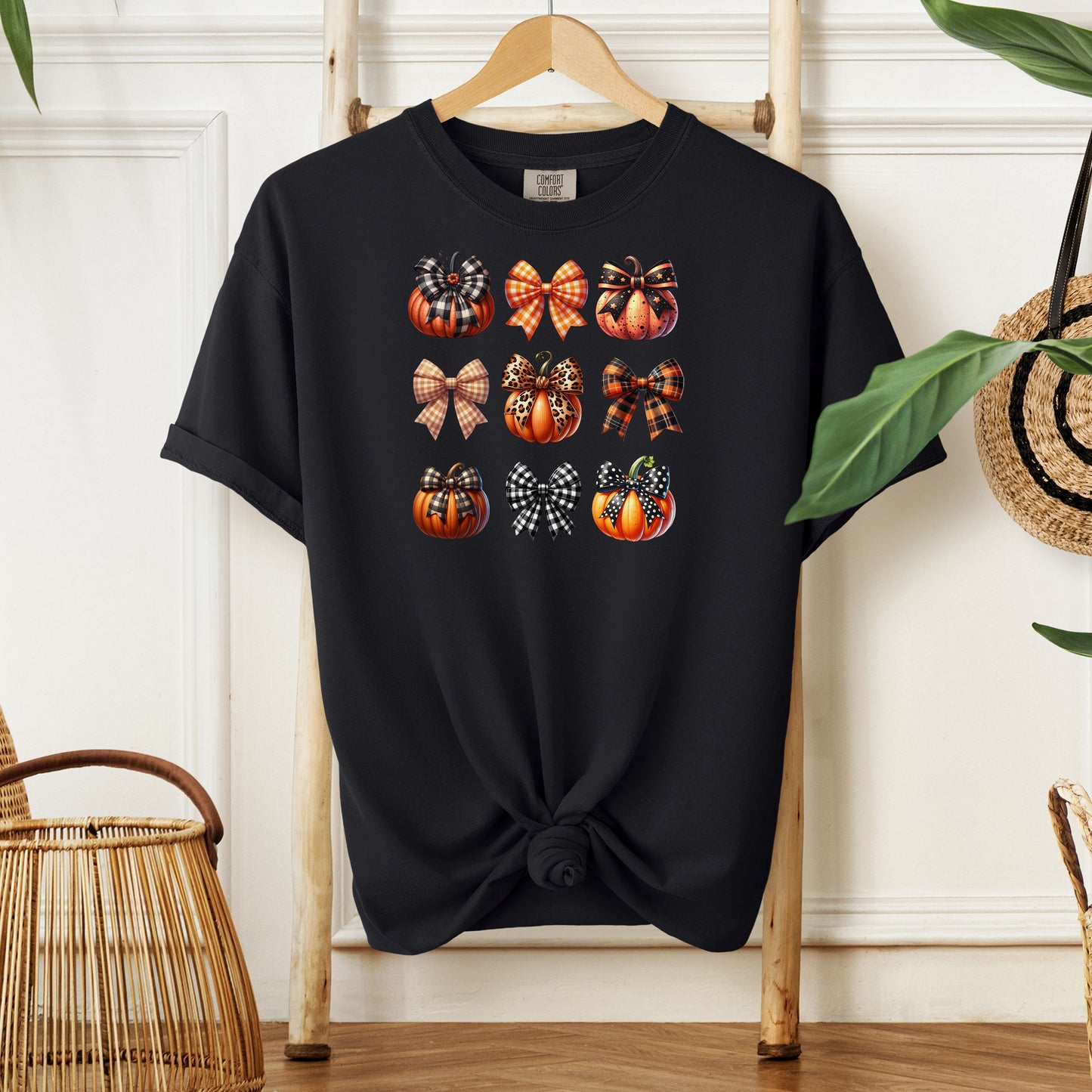 Black T-shirt featuring “Pumpkins and Coquette Bows” design. The artwork displays a grid of pumpkins and decorative coquette bows in plaid, gingham, and fall patterns, blending rustic autumn charm with a playful coquette aesthetic. Perfect for celebrating fall fashion and cozy seasonal vibes with a cute, stylish twist.