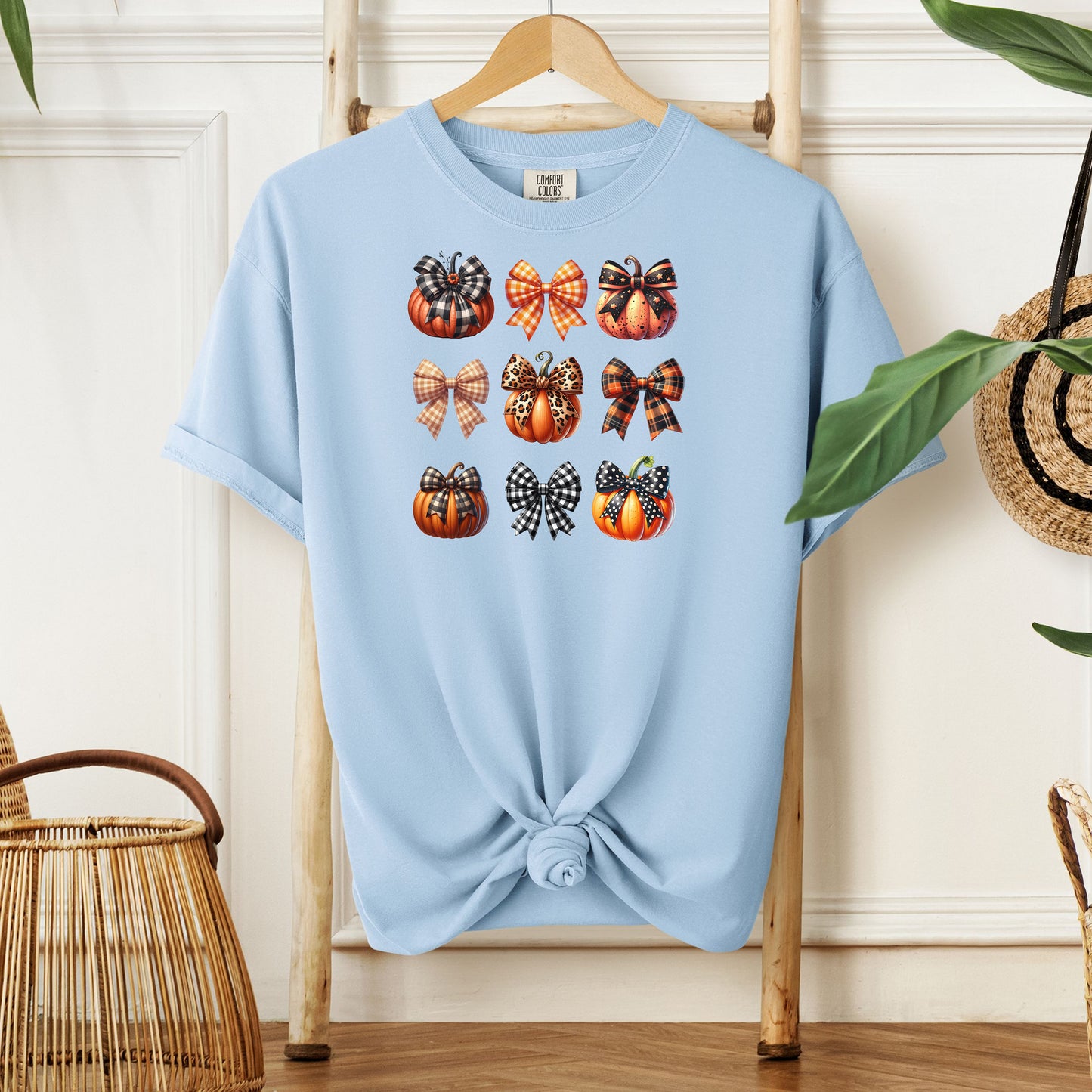 Chambray light blue T-shirt featuring “Pumpkins and Coquette Bows” design. The artwork displays a grid of pumpkins and decorative coquette bows in plaid, gingham, and fall patterns, blending rustic autumn charm with a playful coquette aesthetic. Perfect for celebrating fall fashion and cozy seasonal vibes with a cute, stylish twist.