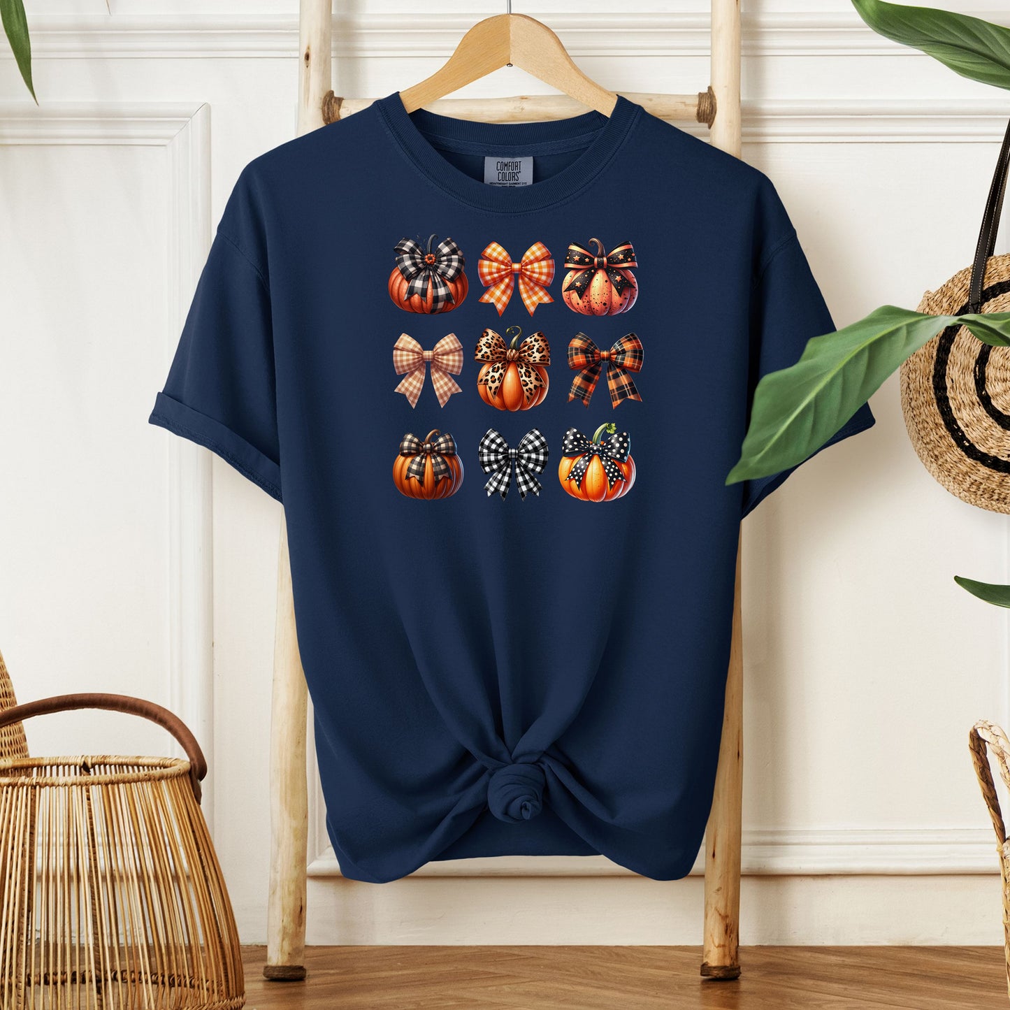 True Navy T-shirt featuring “Pumpkins and Coquette Bows” design. The artwork displays a grid of pumpkins and decorative coquette bows in plaid, gingham, and fall patterns, blending rustic autumn charm with a playful coquette aesthetic. Perfect for celebrating fall fashion and cozy seasonal vibes with a cute, stylish twist.