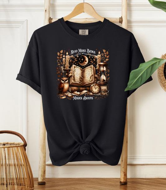 A black t-shirt featuring a whimsical "Read More Books, Magick Awaits" design. The graphic showcases an open enchanted book surrounded by candles, a crescent moon, owl, pocket watch, hourglass, and magical elements, creating a mystical and literary aesthetic perfect for book lovers and fantasy fans.