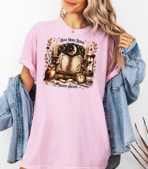 Person wearing a blossom pink t-shirt featuring a whimsical "Read More Books, Magick Awaits" design. The graphic showcases an open enchanted book surrounded by candles, a crescent moon, owl, pocket watch, hourglass, and magical elements, creating a mystical and literary aesthetic perfect for book lovers and fantasy fans.